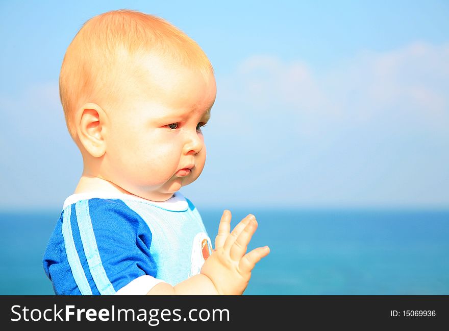 Beautiful white little boy in a bright blue t-shirt is seriously looking into the future put out his hand against the background of summer sea and blue sky. Beautiful white little boy in a bright blue t-shirt is seriously looking into the future put out his hand against the background of summer sea and blue sky