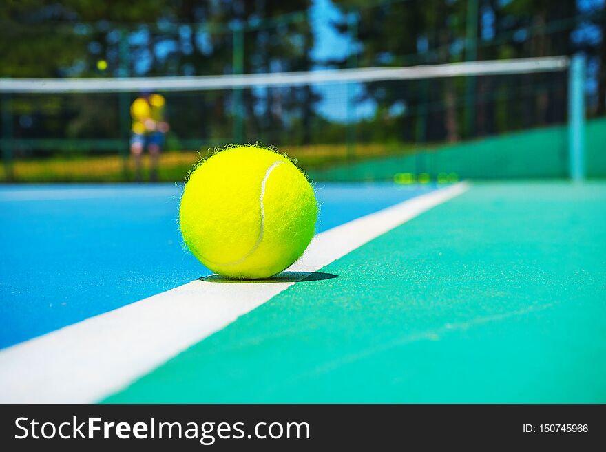 Tennis ball on white court line on hard modern blue green court with player, net, balls, trees on the background. Close-up, selective focus. Sport, tennis play, healthy lifestyle concept.