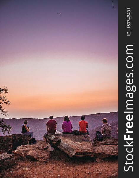 A group of young travelers sitting on top of a mountain and watching a beautiful view of the canyon and the sunset sky