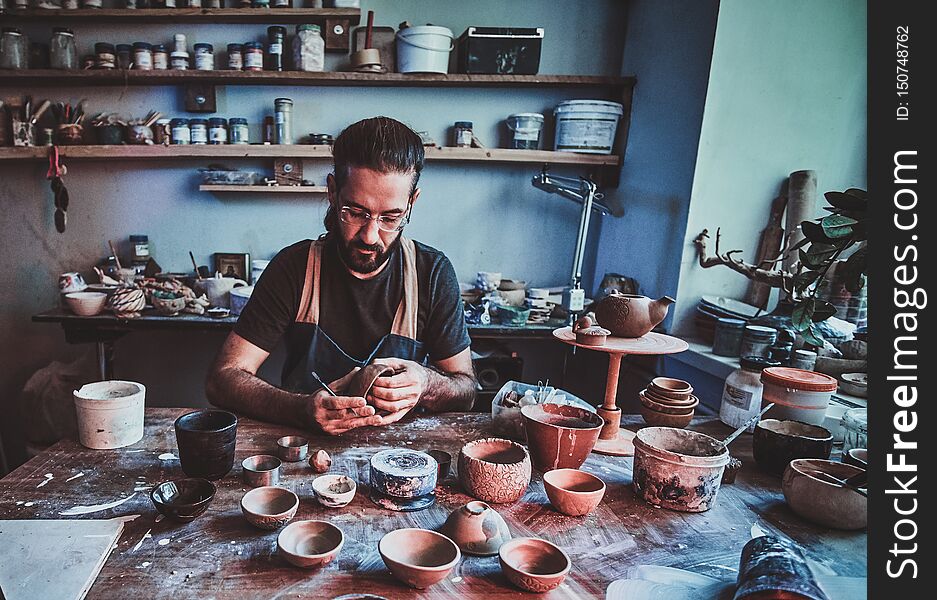 Pensive entusiastici potter in glasses at his own workshop is workig on a new handmade teapot.