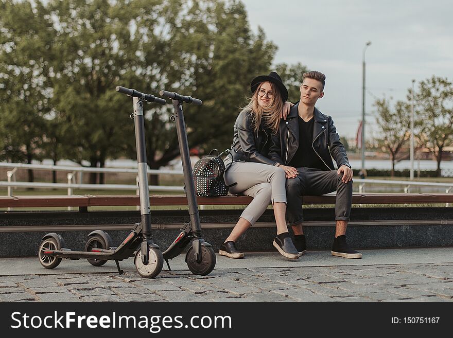 Happy trendy couple is chilling on the bench at city park with their scooters. Happy trendy couple is chilling on the bench at city park with their scooters.