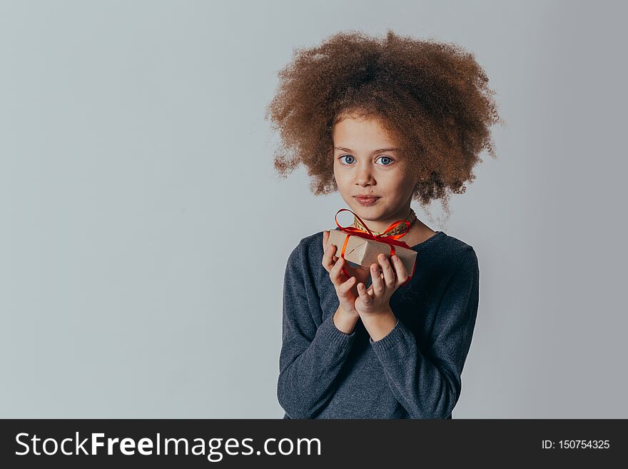 Portrait Of A Smiling Cute Girl With Curly Hair And A Red Gift In Her Hands. Studio Shot