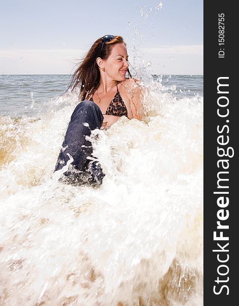 Young adult woman in wet jeans sits in splashes of tidal waves of a sea smiling. Young adult woman in wet jeans sits in splashes of tidal waves of a sea smiling