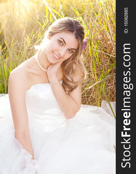 An attractive young bride wearing a white wedding dress and pearls is sitting with her head on her hand in the grass. Vertical shot. An attractive young bride wearing a white wedding dress and pearls is sitting with her head on her hand in the grass. Vertical shot.