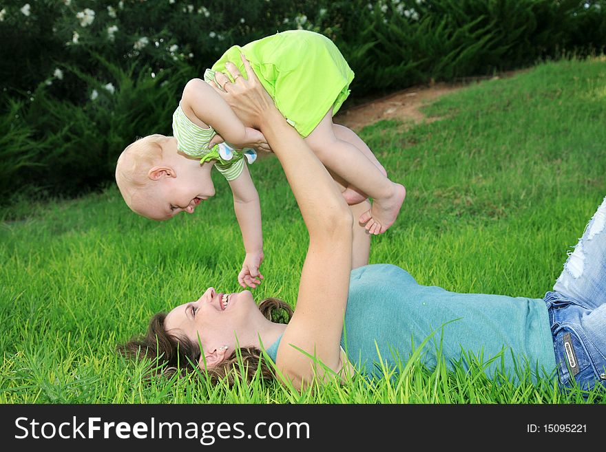 Happy young mother lying on green grass and smiling at her little son lifted him up