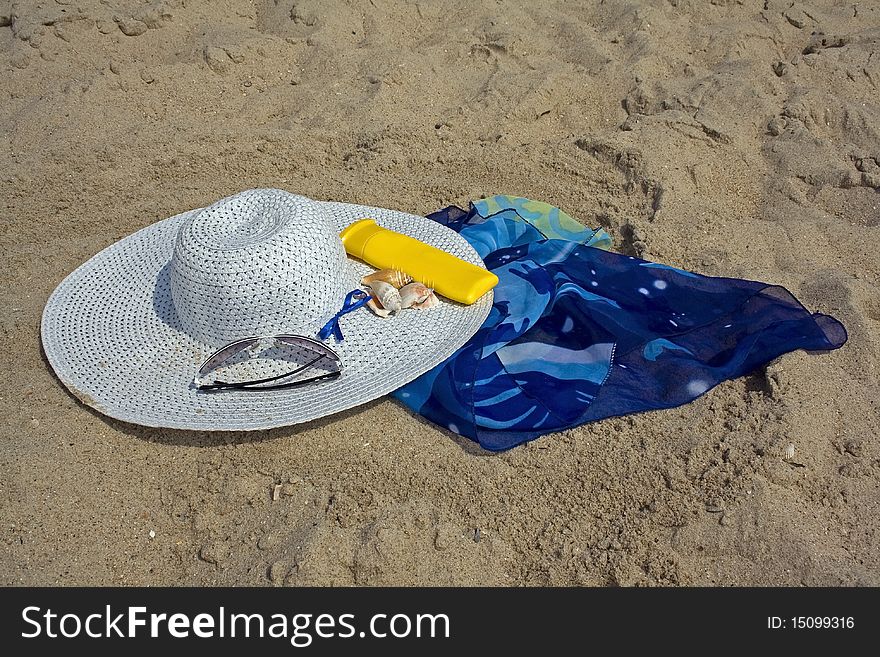 A hat, sunglasses, cape, suntan cream, clams on the background of beach sand. A hat, sunglasses, cape, suntan cream, clams on the background of beach sand