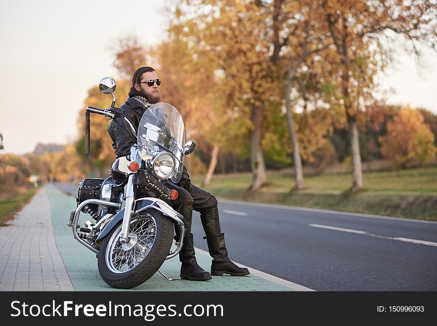 Young bearded biker in black leather clothing and dark sunglasses sitting on motorcycle on clean paved roadside, on background of empty straight asphalt road and vintage trees golden bokeh foliage. Young bearded biker in black leather clothing and dark sunglasses sitting on motorcycle on clean paved roadside, on background of empty straight asphalt road and vintage trees golden bokeh foliage.