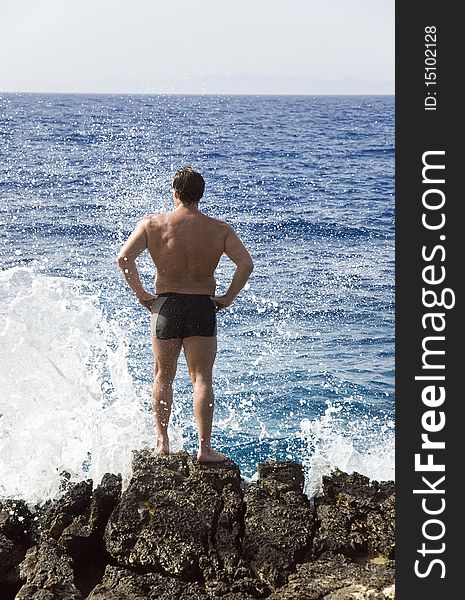 A color portrait photo of a fit looking caucasian male standing on some rocks while a wave breaks in front of him in Paxos Greece. A color portrait photo of a fit looking caucasian male standing on some rocks while a wave breaks in front of him in Paxos Greece.