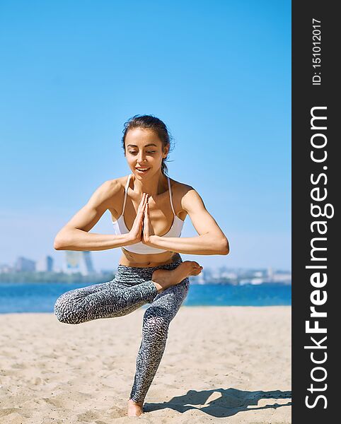 Young Slim Athletic Woman Makes Yoga And Balance Exercises On The Sand Beach With City Background. Healthy Lifestyle