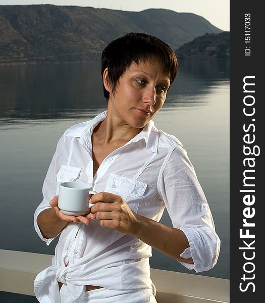 Young dressed white brunette woman meets sunrise drinking morning coffee on the mountain and sea background