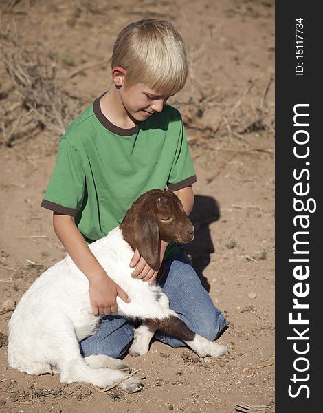 A boy sitting in a pasture holding on to a new baby goat, with a small smile on his face. A boy sitting in a pasture holding on to a new baby goat, with a small smile on his face.