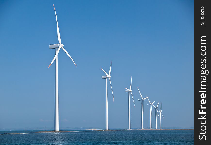 Seven windmills lined up on a dam reaching out from the coast with clear blue sky. Seven windmills lined up on a dam reaching out from the coast with clear blue sky.