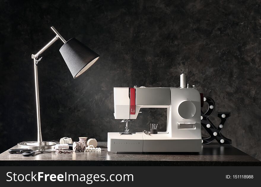 Modern sewing machine with tailor's supplies and lamp on table near dark wall