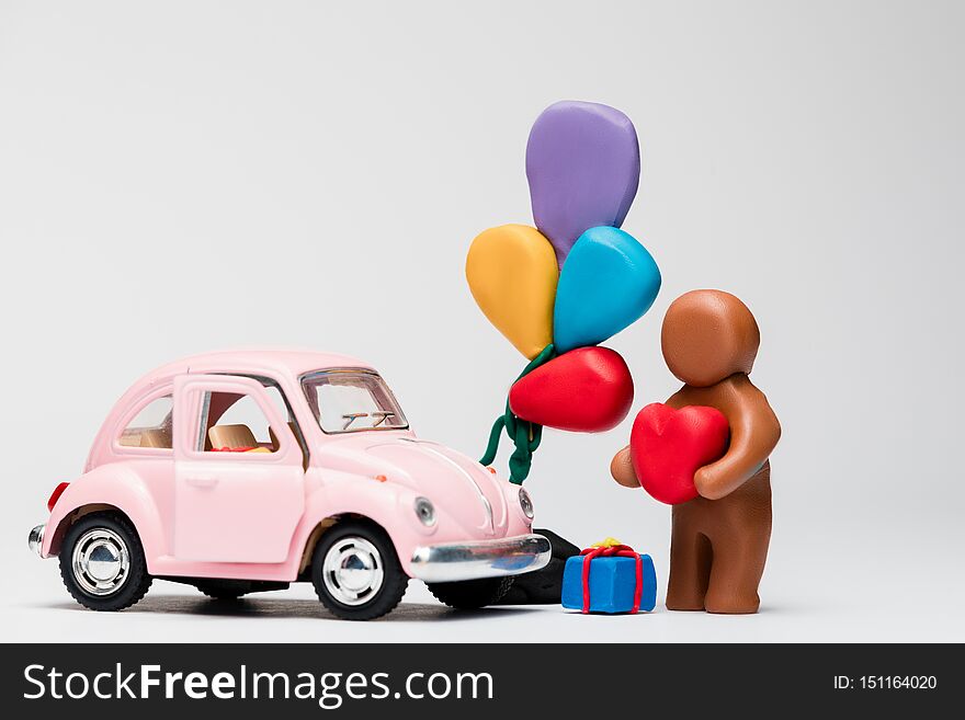 A man made from brown plasticine next to a pink car with balloons on white background, aligned in the center