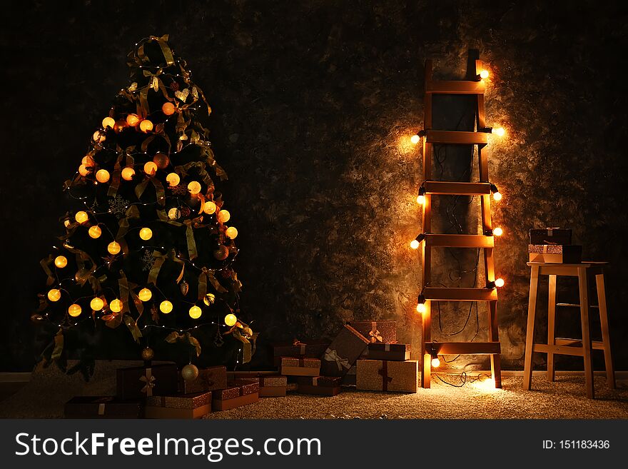 Stylish Christmas fir tree with garland and gifts near dark wall indoors