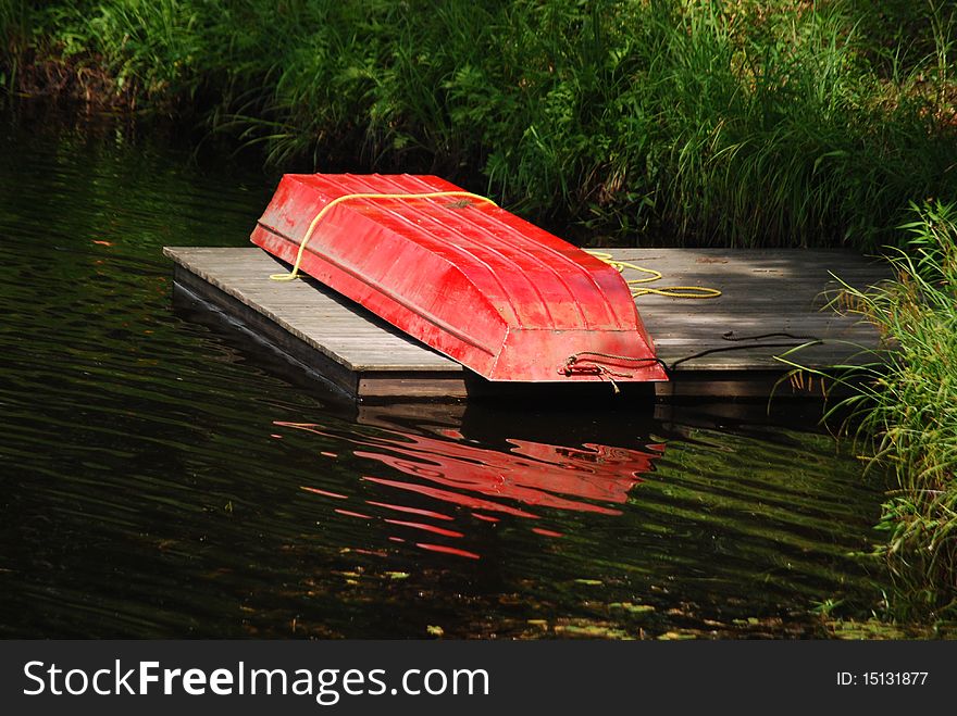 A red small row boat out of water upside down on a dock in a pond. A red small row boat out of water upside down on a dock in a pond.