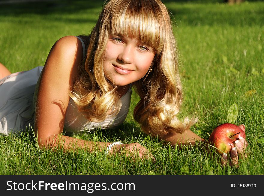 A beautiful young blonde with an apple lies on a grass in a city park