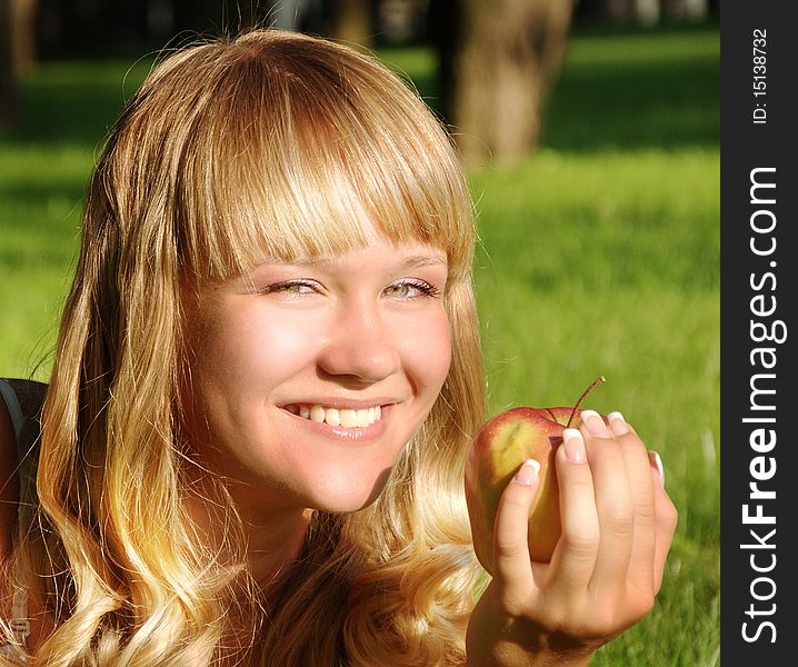 A beautiful young blonde with an apple lies on a grass in a city park