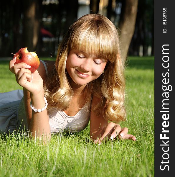 A beautiful young blonde with an apple lies on a grass in a city park