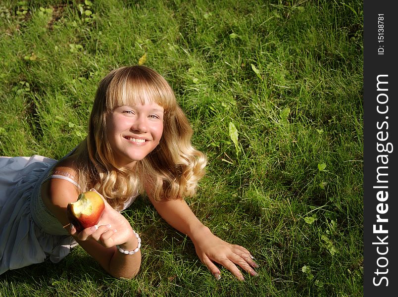 A beautiful young blonde with an apple lies on a grass in a city park