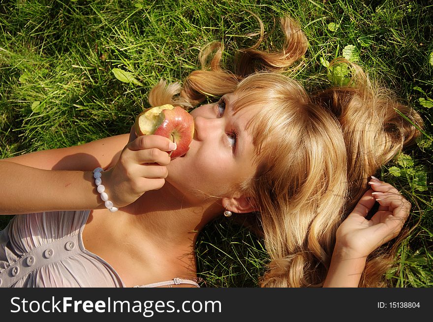 A beautiful young blonde with an apple lies on a grass in a city park