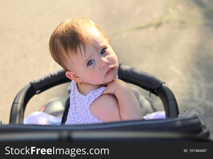 little, beautiful, smiling, cute redhead baby in a sleeveless shirt in a pram out-of-doors drops hands down and looking back