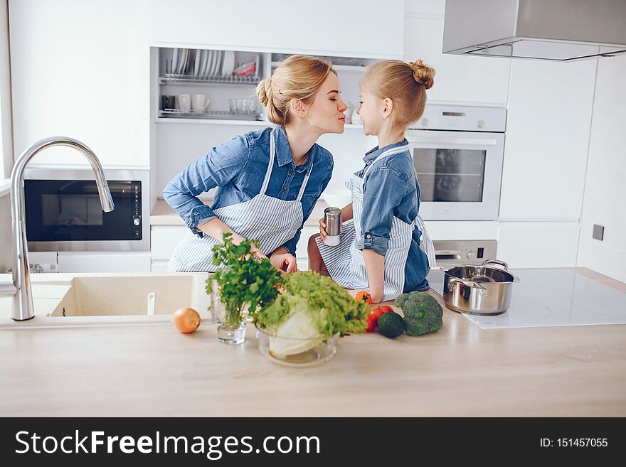A young and beautiful mother in a blue shirt and apron is preparing a fresh vegetable salad at home in the kitchen, along with her little cute daughters with light hair. A young and beautiful mother in a blue shirt and apron is preparing a fresh vegetable salad at home in the kitchen, along with her little cute daughters with light hair