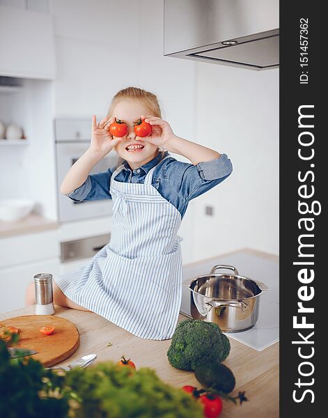 A little and beautiful girl in a blue shirt and apron is preparing a fresh vegetable salad at home in the kitchen. A little and beautiful girl in a blue shirt and apron is preparing a fresh vegetable salad at home in the kitchen