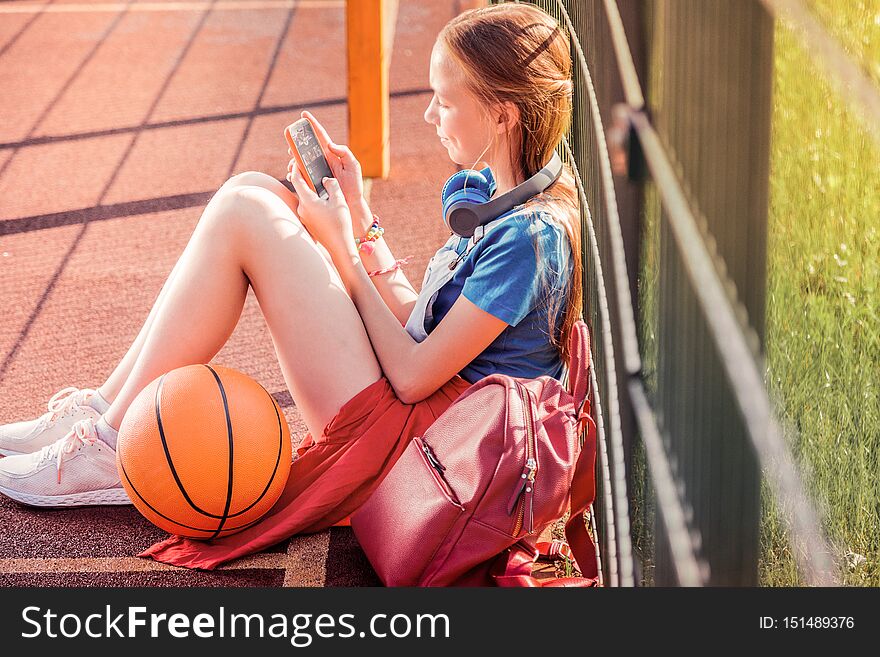 Holidays at school. Focused long-haired girl sitting on a basketball playground and chatting on a smartphone