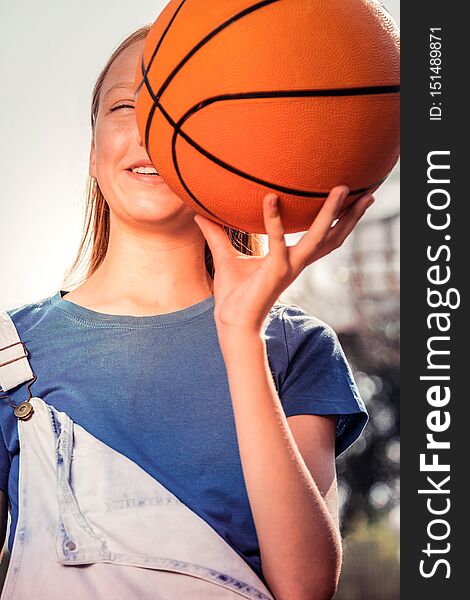 Holding basketball ball . Cheerful young sportive lady in blue t-shirt carrying ball near her face while having fun in a park