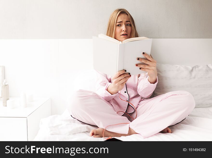 Portrait of young woman sitting on bed and amazedly looking in book at home