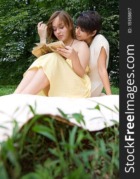 Two young girlfriends reading a book sitting on the coverlet outdoors. Two young girlfriends reading a book sitting on the coverlet outdoors