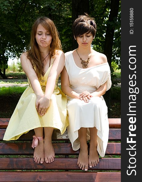 Two attractive young girls sitting on the bench in the park making funny facial expressions. Two attractive young girls sitting on the bench in the park making funny facial expressions