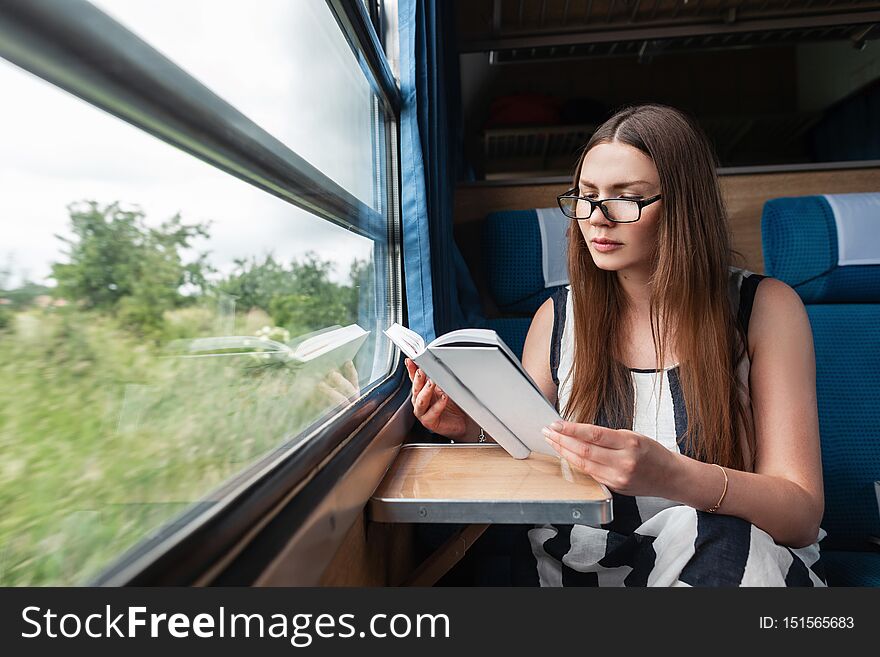 Pretty young woman in a stylish striped summer dress in glasses sits in the train near the window and reads an interesting book.