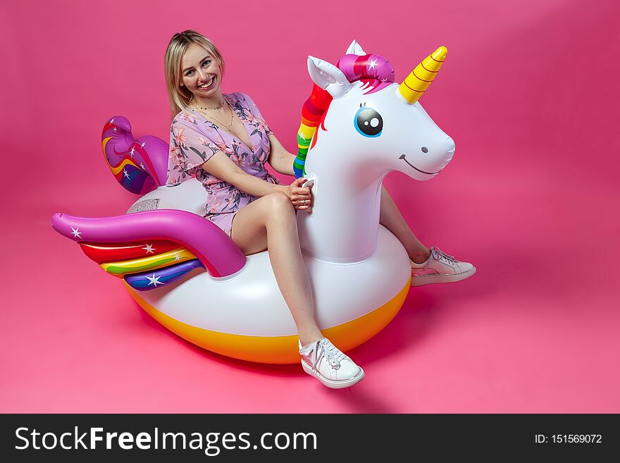 A beautiful blonde girl in a sexy sundress with slim legs in white sneakers sits on an inflatable multi-colored unicorn on a pink