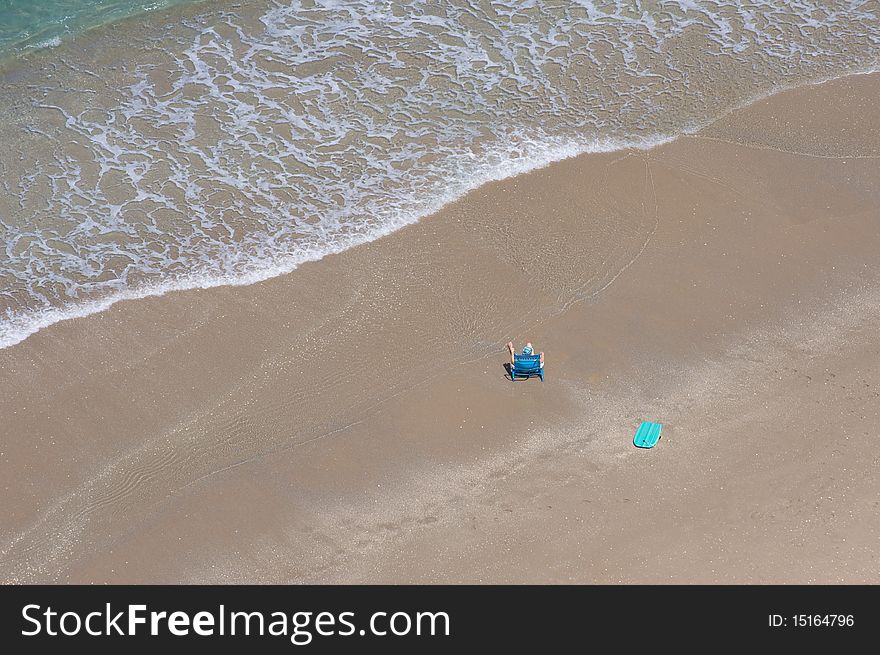 Man alone at the beach on a beach chair. There is a boogie board near him. The ocean is tranquil