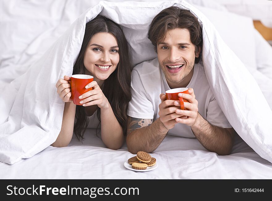 Good morning coffee. Cheerful couple hiding under duvet with cups and cookies, free space