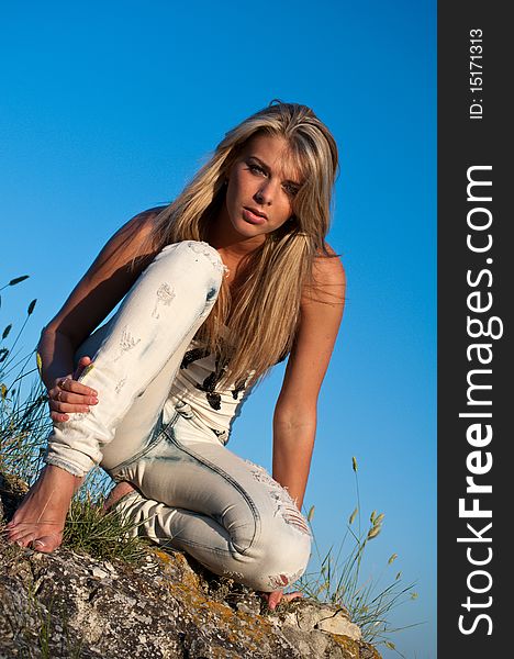 Barefoot blonde young woman sitting on a rock against the blue summer sky