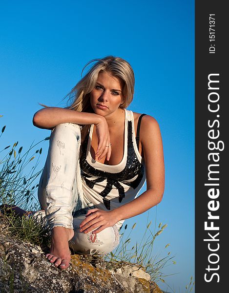 Barefoot blonde young woman sitting on a rock against the blue summer sky