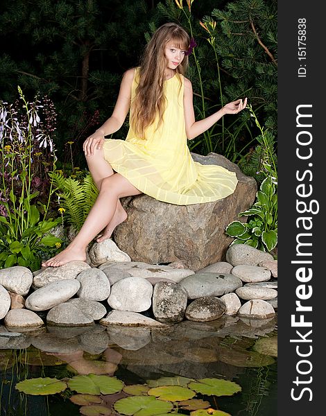 The girl in a yellow dress sits on the big stone near a pond