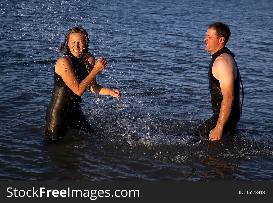 A couple in their wet suits playing and splashing around while they are standing in the water. A couple in their wet suits playing and splashing around while they are standing in the water.