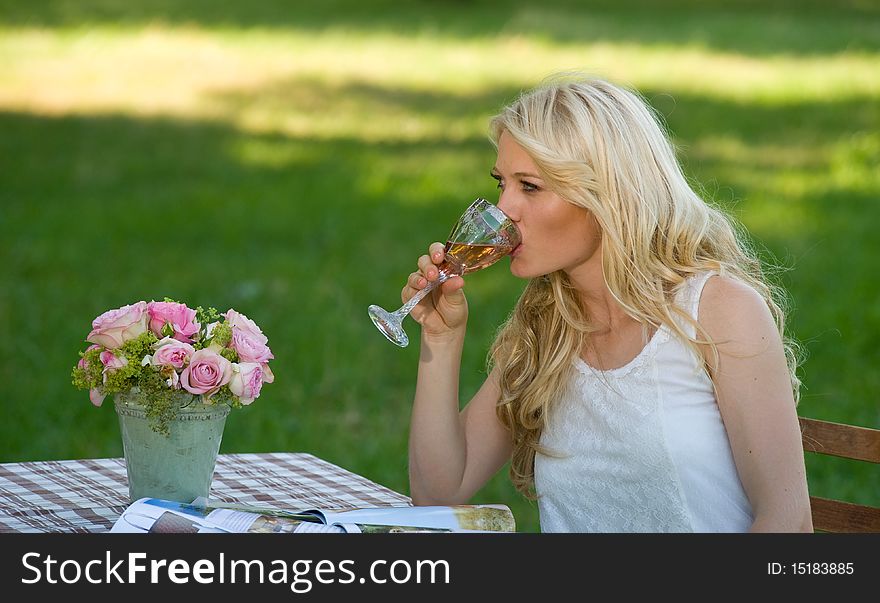Young attractive blond with a glass of wine in her hand sitting in the green park on a folding table. Young attractive blond with a glass of wine in her hand sitting in the green park on a folding table