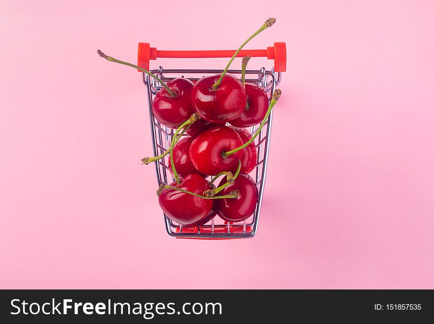 Creative top view of ripe cherries in a shopping cart on pink  background in minimal style. Concept of supermarket or fruit market shopping