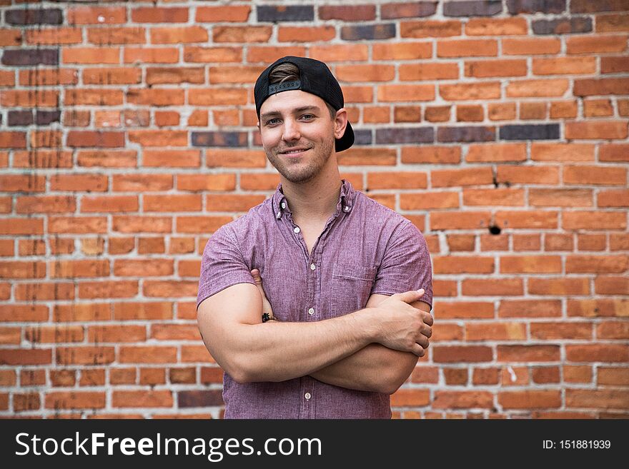 Handsome Young Caucasian Man With Cellphone And Backwards Hat Smiling For Portraits In Front Of Textured Brick Wall Outside