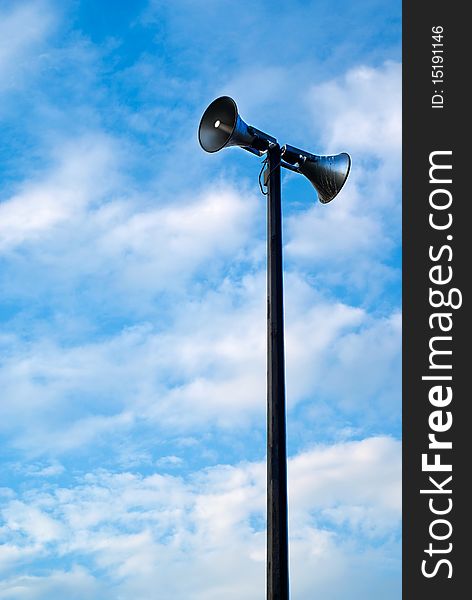 A public address system speakers tower against a cloudy, blue, daytime sky. A public address system speakers tower against a cloudy, blue, daytime sky.