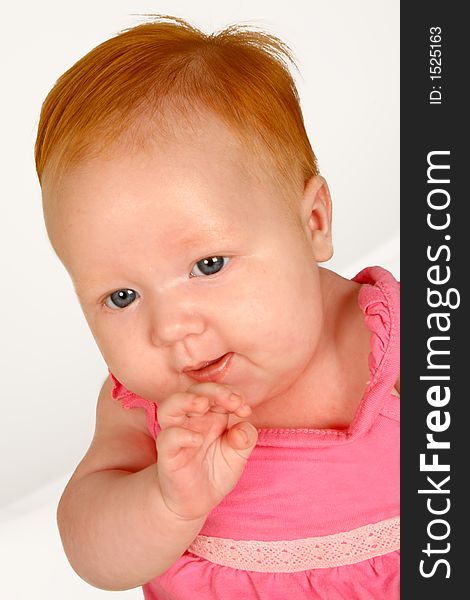 A baby girl sitting holding her hand by her mouth. A baby girl sitting holding her hand by her mouth