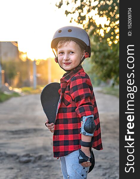 Happy little boy standing on the road holding a skate with his hands. the child defended himself, he put on hand safety gloves elbows and knees wearing protective shields. on the head wearing a helmet