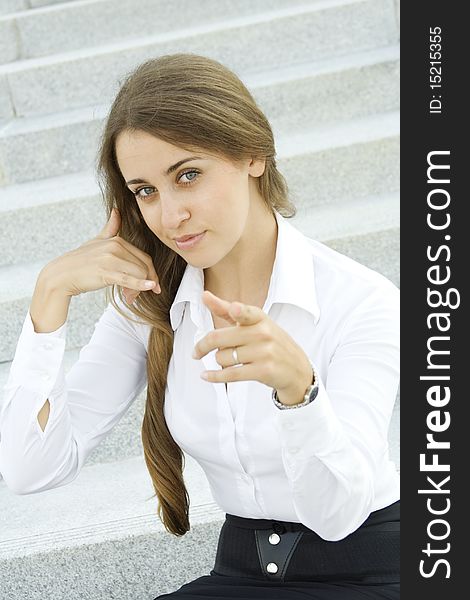 Young businesswoman outdoors on the stairs near the office building shows a sign of Call me pointing to viewer. Young businesswoman outdoors on the stairs near the office building shows a sign of Call me pointing to viewer