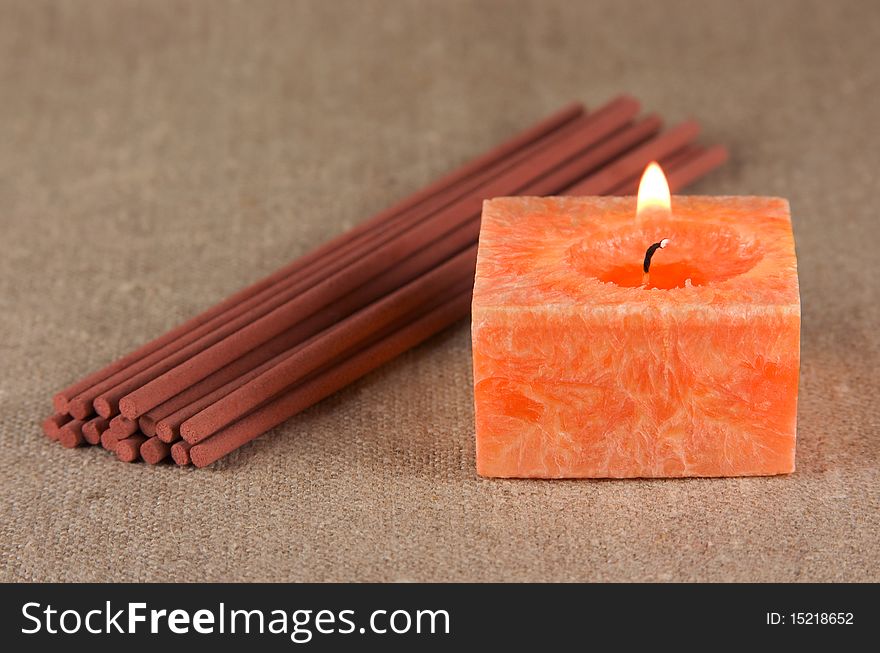 A bundle of incense sticks and orange burning candle on the fabric background. A bundle of incense sticks and orange burning candle on the fabric background