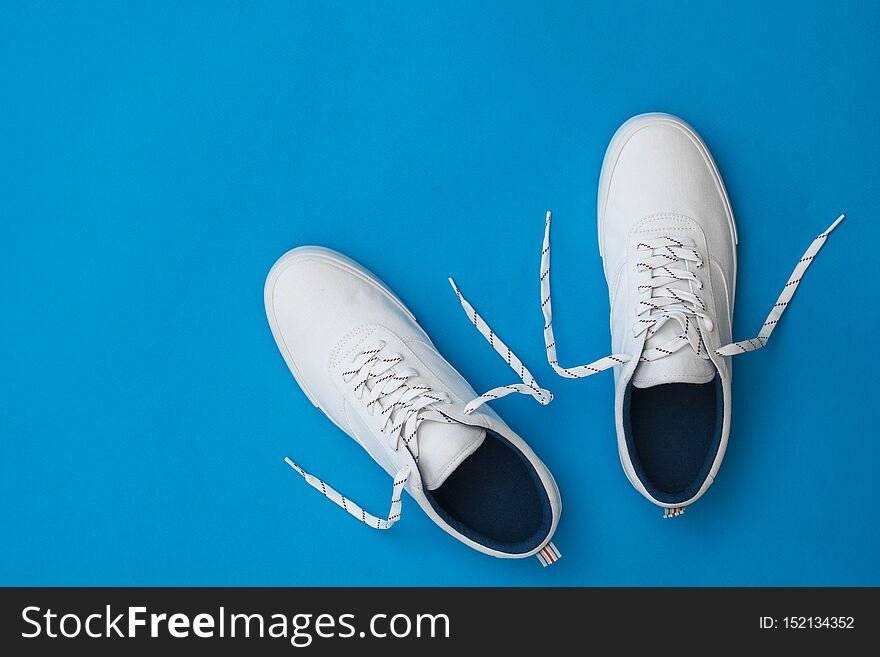 White sneakers with untied laces on a blue background. Sports style. Flat lay. The view from the top.